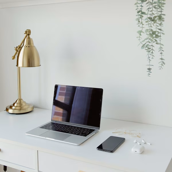 A neatly arranged white office desk with a laptop, brass task lamp, and greenery